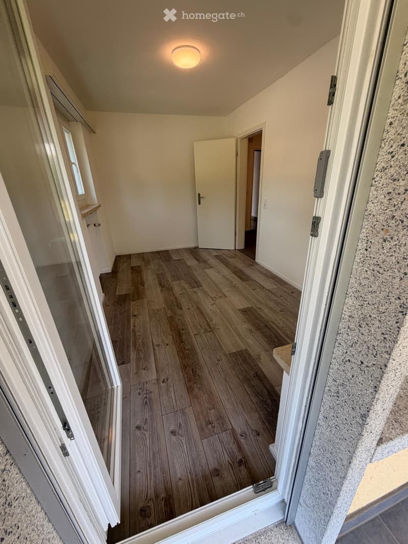 Empty hallway with wooden flooring, white walls, and a ceiling light. Features an open door leading to another room and a window on the left side.