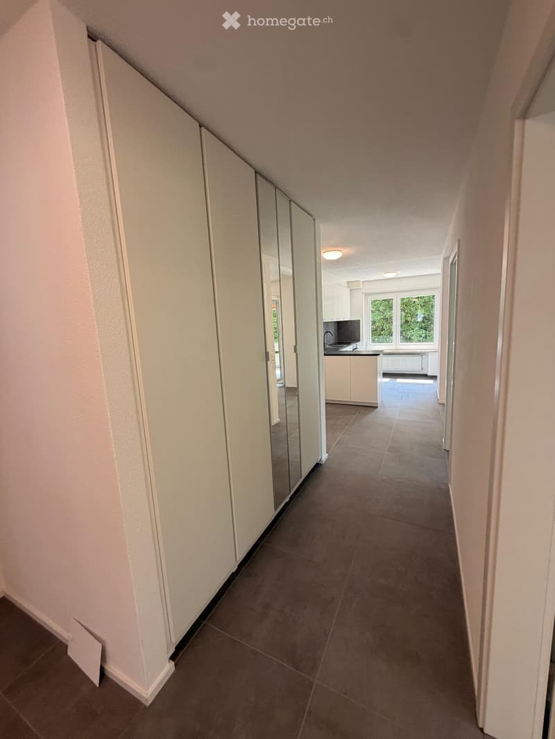 A clean, modern hallway with tiled flooring, white walls, and a mirrored wardrobe on one side. The hallway leads to a kitchen area visible at the end, featuring white cabinets, a sink, and windows with greenery outside.