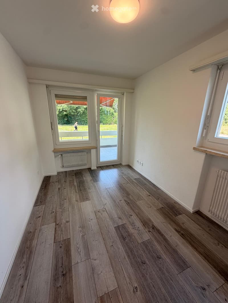 Empty room with wooden flooring, white walls, large windows, and a sliding glass door leading to an outdoor area.