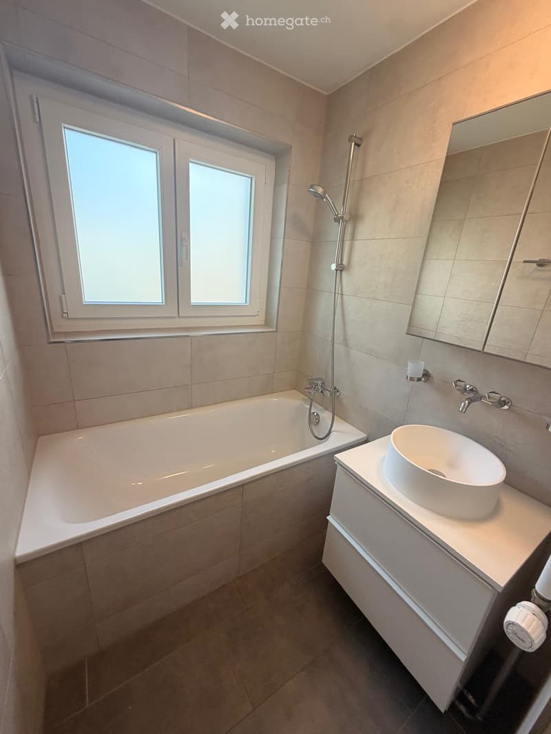 Modern bathroom with a bathtub, sink, mirror, and showerhead. Features tiled walls and a window with frosted glass panes.