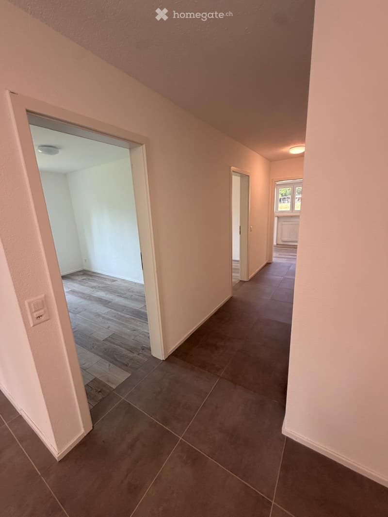 Empty hallway with tiled floor and wooden flooring in adjacent room, featuring neutral-colored walls and recessed lighting.