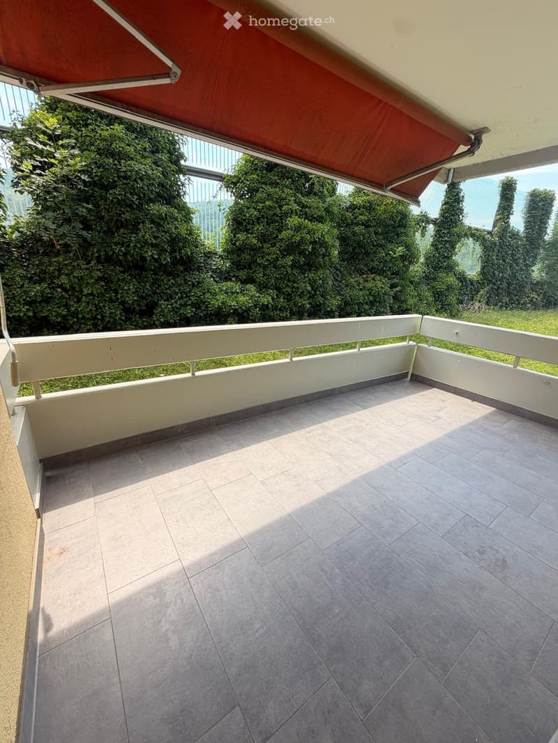 Balcony with tiled floor, white railing, and a red awning providing shade. Greenery visible in the background.