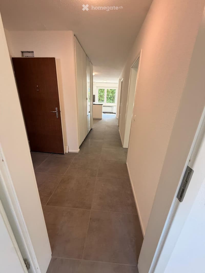 A long, narrow hallway with tiled flooring and white walls, featuring several doors and a glimpse of a room at the end with windows and furniture.