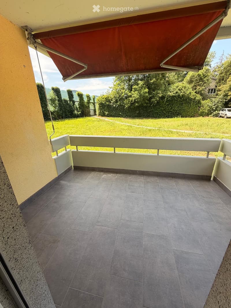 Balcony with tiled floor, white railing, and a red awning providing shade. The view includes a green lawn and trees.