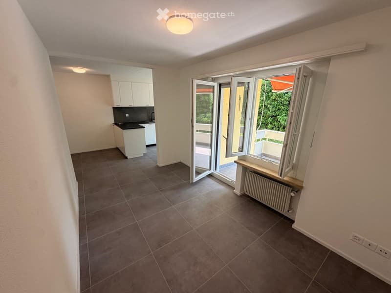 Empty room with tiled floor, white walls, and a large window opening to a balcony. A small kitchen area is visible in the background with white cabinets and a countertop.