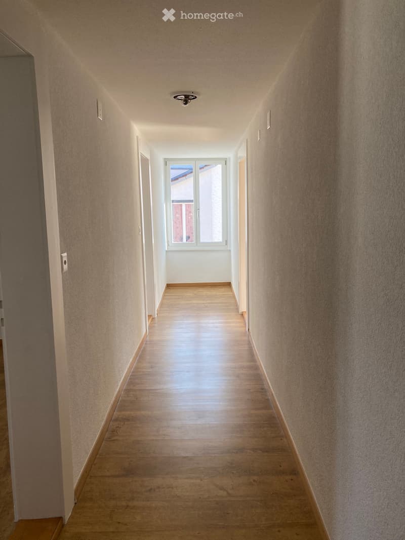 Empty hallway with wooden flooring, beige walls, and a window at the end allowing natural light to enter.