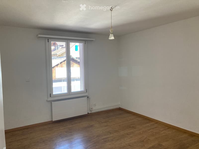 Empty room with wooden flooring, white walls, a window with white frame, radiator, and a hanging light fixture.