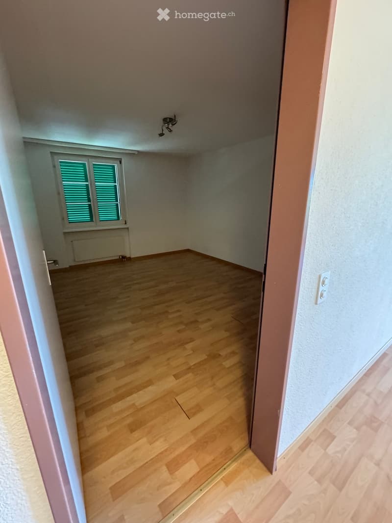 Empty room with wooden flooring, white walls, and a window with green shutters. No furniture or equipment is present.