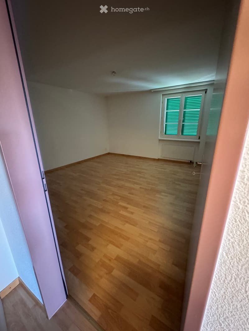 Empty room with wooden flooring, white walls, and a window with green shutters. No furniture or equipment is present.