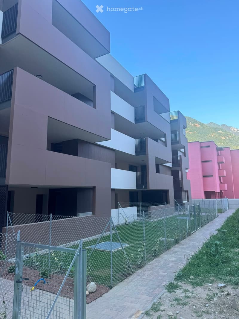 Modern multi-story residential building with balconies, surrounded by a fenced area and pathway.