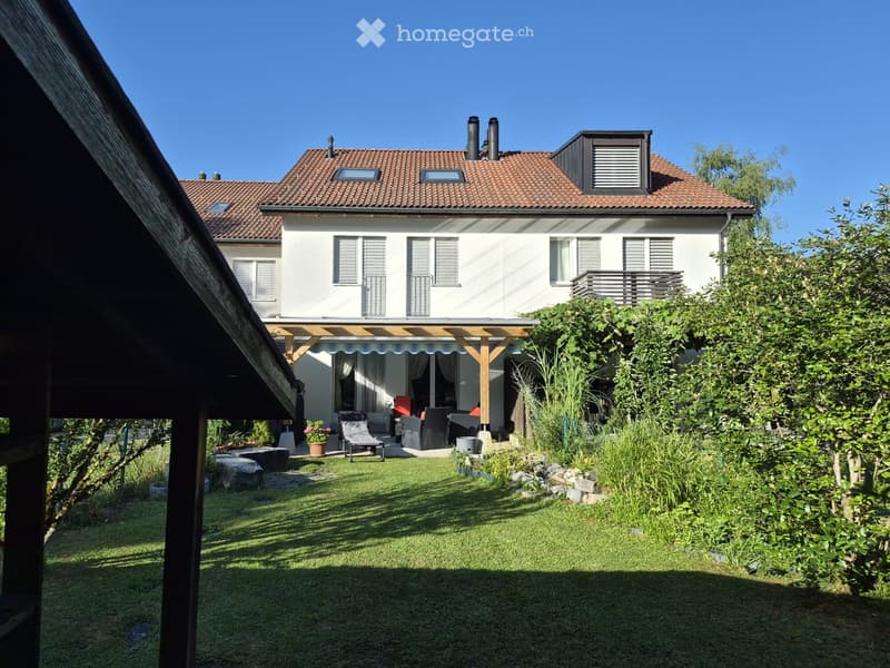 Garden area with a wooden pergola, lounge chairs, a table, and a flower pot, surrounded by greenery and a well-maintained lawn.