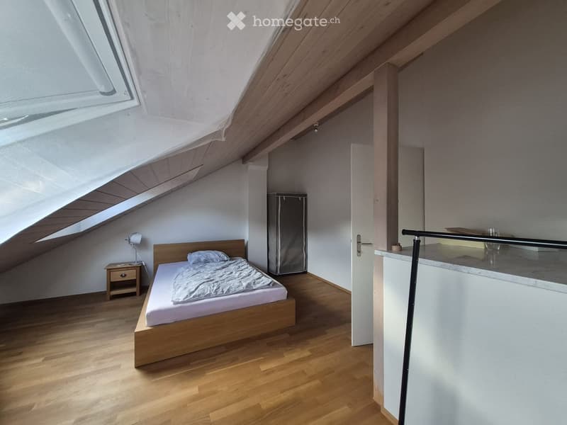 Modern attic bedroom with wooden flooring, slanted wooden ceiling, skylight window, bed with white bedding, nightstand with lamp, and a small refrigerator.