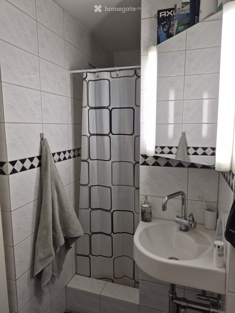 A small bathroom with white tiled walls and floor, featuring a sink with a faucet, a mirror, a towel hanging on the wall, and a shower area with a black and white patterned shower curtain.