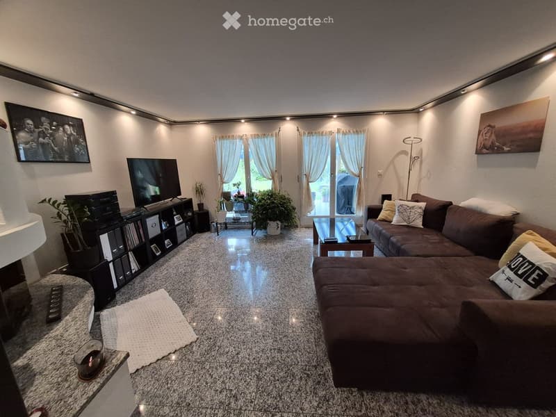 Modern living room with a dark brown sectional sofa, coffee table, TV, bookshelf, and decorative plants. Large windows with sheer curtains lead to a balcony.