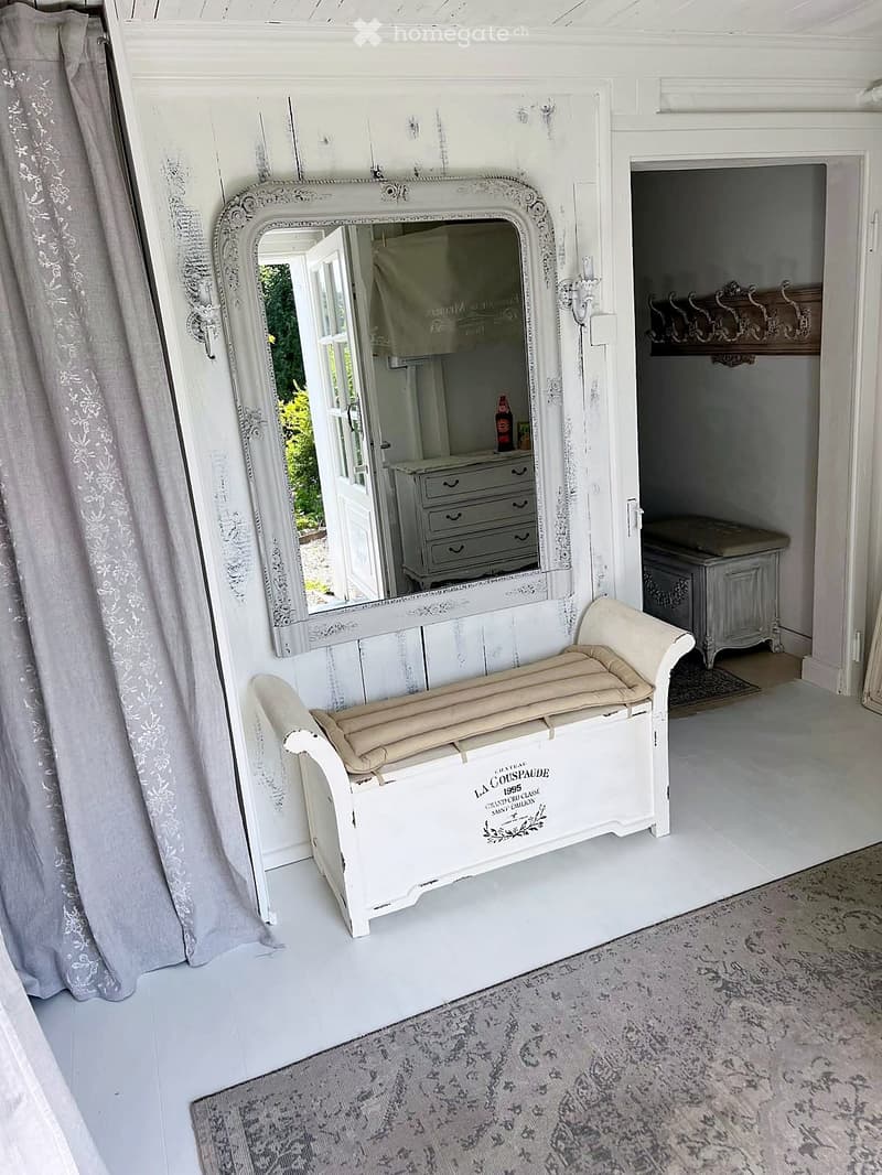 Furnished Room. A well-lit, elegantly furnished room with a white bench, ornate mirror, and a small chest of drawers visible through an open doorway.