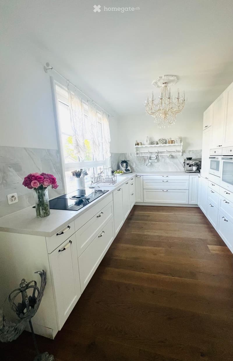 Bright kitchen with white cabinets, wooden flooring, and a chandelier. Features a sink, stove, microwave, coffee maker, and a vase with pink flowers on the counter.