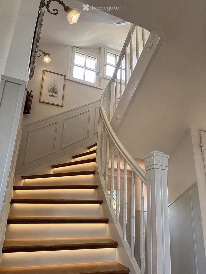 Spiral staircase with wooden steps and white balustrade, illuminated by integrated step lighting and wall-mounted lamps, with a framed artwork and a fire extinguisher on the wall.