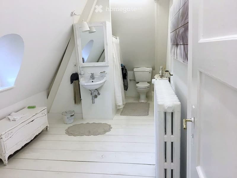Bathroom. Cozy attic bathroom with white wooden floors, a sink, toilet, mirror, radiator, and a small stool with towels.