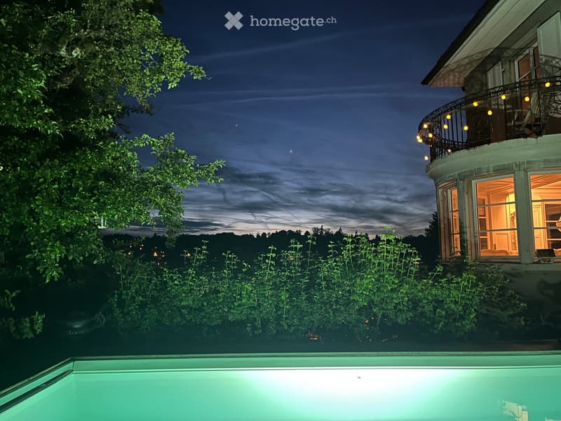 Nighttime view of a swimming pool with a house in the background, featuring a balcony with string lights and a view of the surrounding landscape.