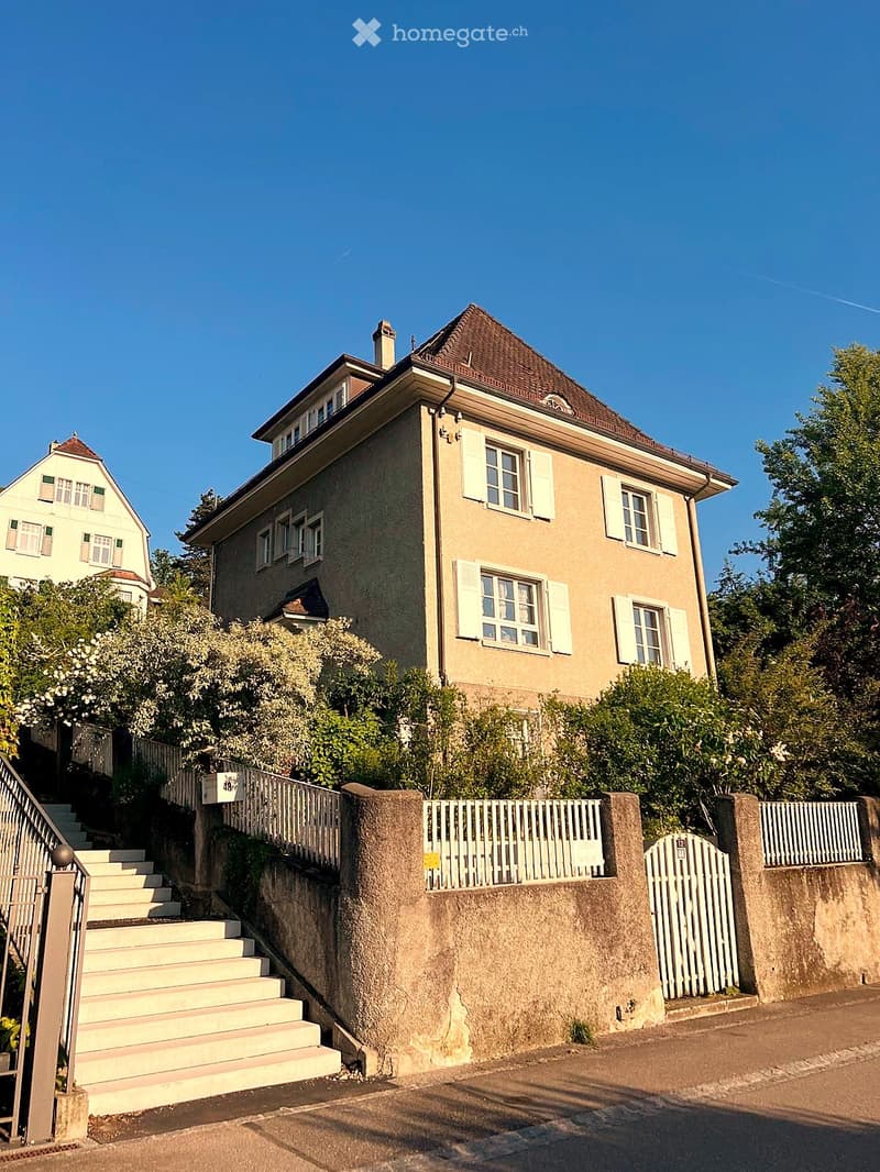 House. Two-story house with beige facade, white shutters, and a sloped roof. The house is surrounded by greenery and accessed via a set of concrete steps leading to a white picket fence.