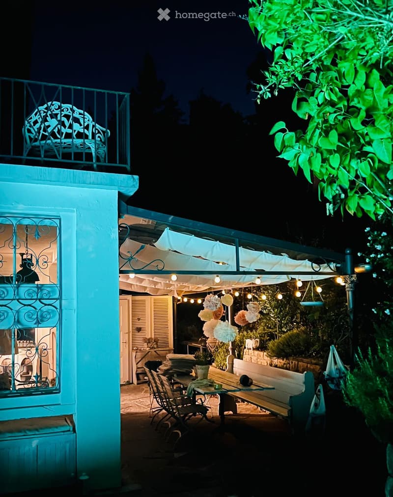 A covered seating area with a table, chairs, and string lights attached to a pergola. It is part of a blue-painted building with a window and a chair on its balcony. The area is outdoors with greenery around it and offers a view of the night sky.