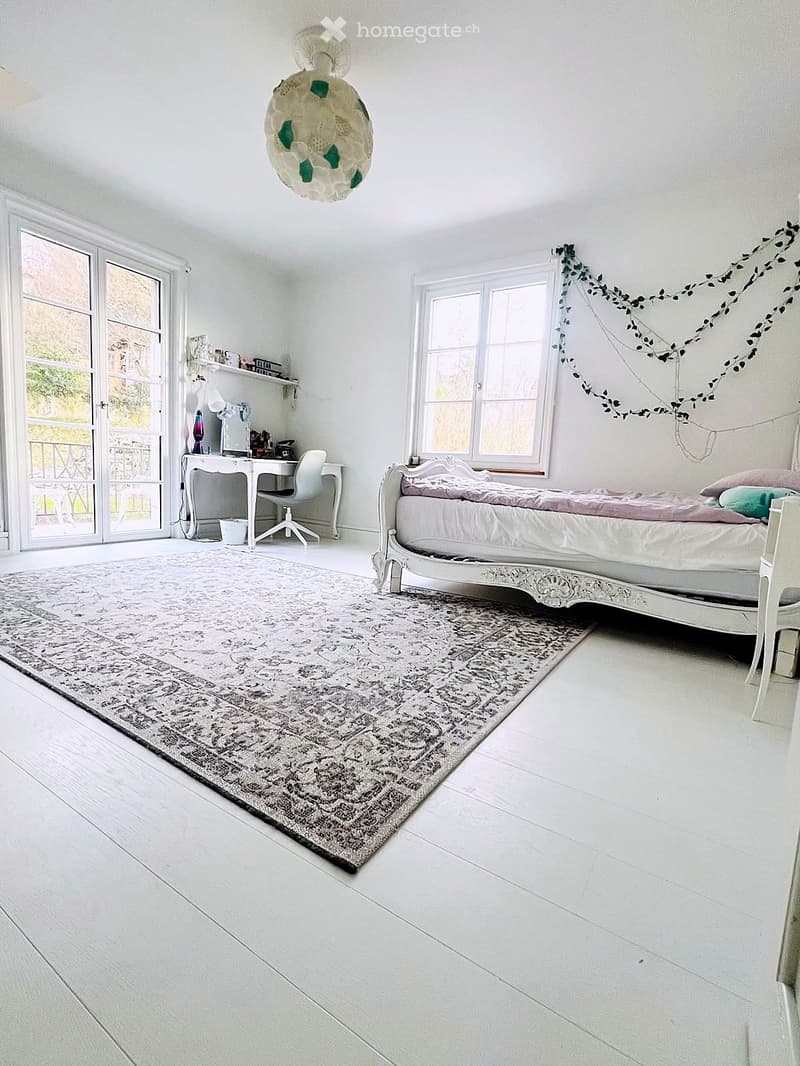 Bedroom. Bright bedroom with white walls, wooden floor, large windows, a white ornate bed, a desk with chair, and a patterned rug.