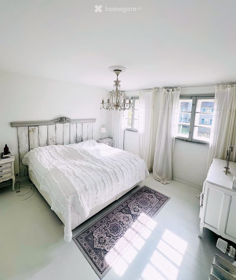 Bedroom. Bright bedroom with white walls, wooden floor, large windows with white curtains, a wooden bed with a white quilt, a chandelier, a nightstand with a lamp, and a patterned rug.