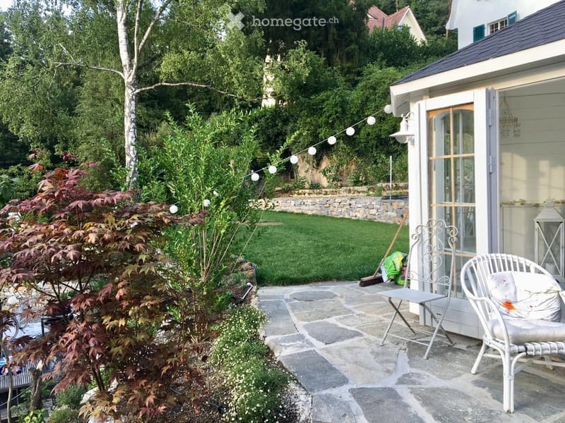 Stone path, white outdoor furniture, hanging string lights, greenery and trees, a small wall, a yard with grassy lawn, a small house on the right side.