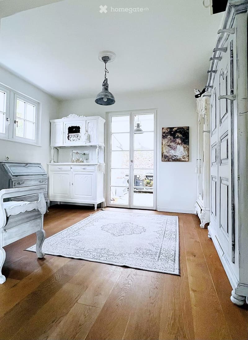 Furnished Room. Bright, airy room with wooden flooring, white walls, and large windows. Features a white cabinet, a vintage chair, a large rug, and a painting on the wall.