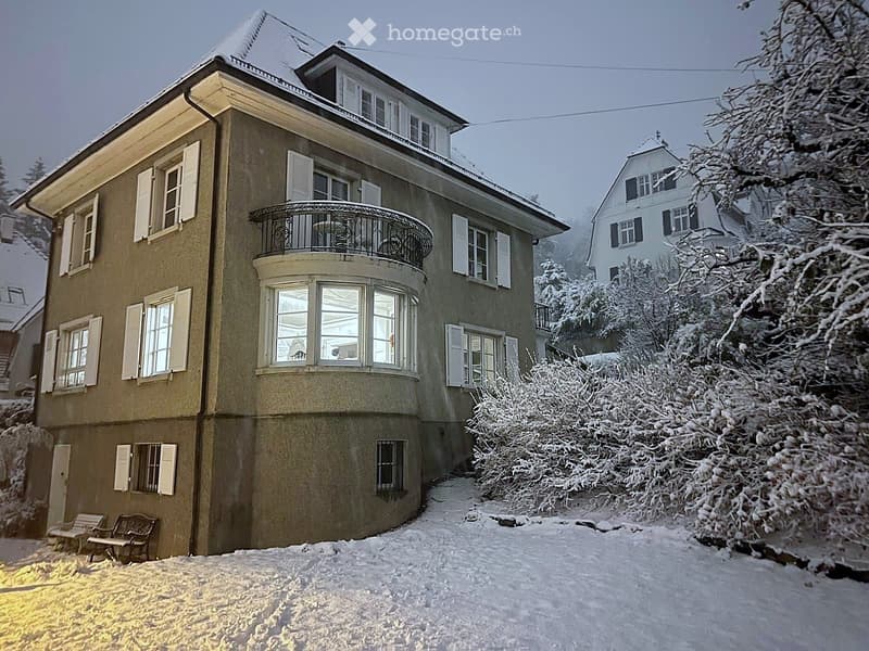 House. A two-story house with beige walls, white shutters, and a balcony on the upper floor, surrounded by snow-covered trees and bushes.