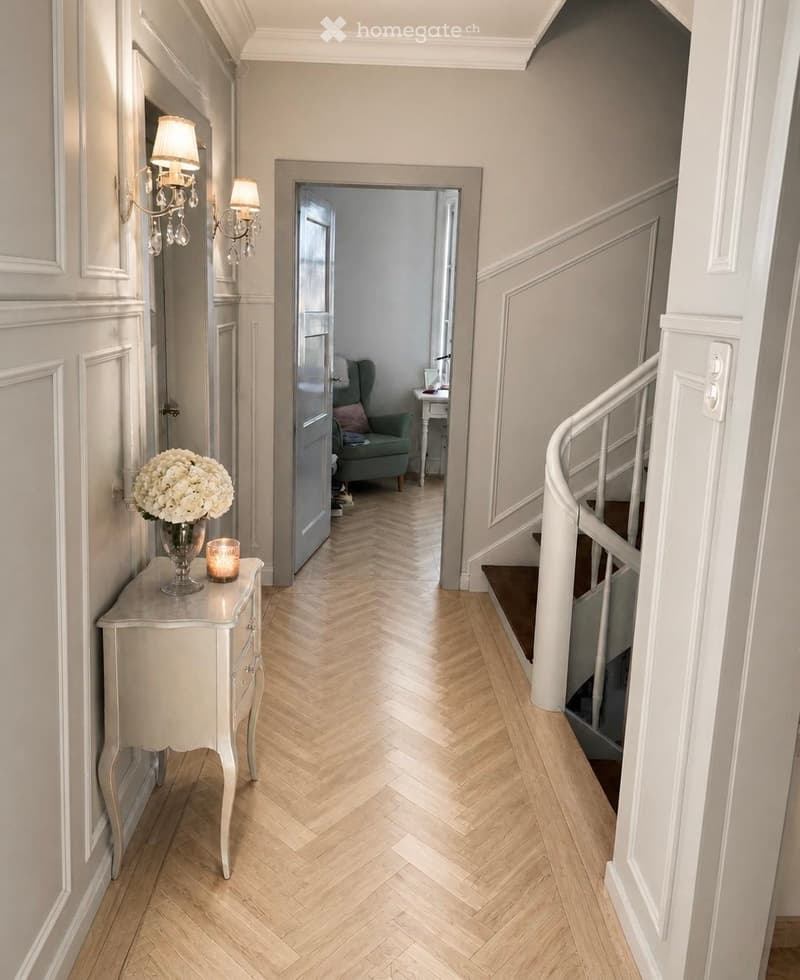 Elegant hallway with herringbone wooden flooring, white walls, decorative side table with vase and candle, wall-mounted chandelier, and a view into a green armchair in the adjacent room.