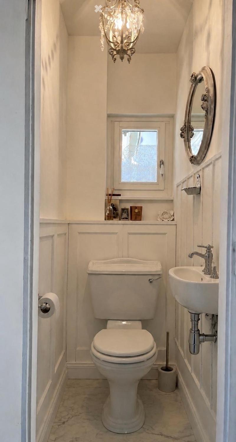 A compact bathroom with white walls, a toilet, a sink with a modern faucet, a round mirror, a toilet paper holder, a toilet brush holder, a small window, and a decorative chandelier.