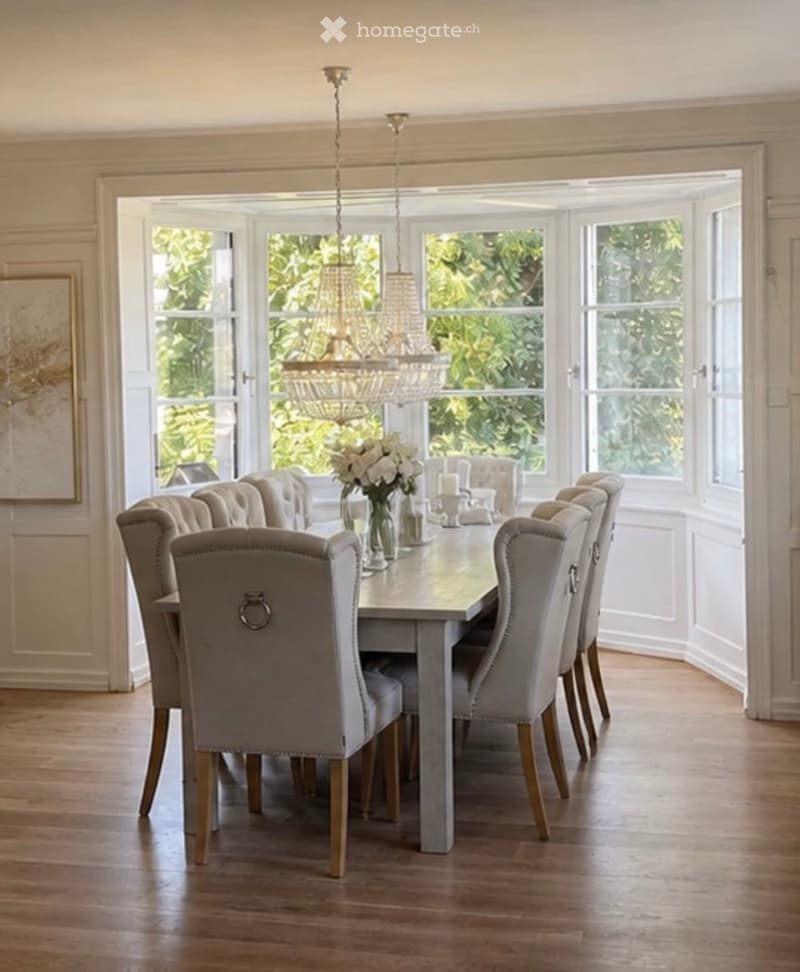 Elegant dining room with a grey dining table, upholstered chairs, a chandelier, and a vase of white flowers on the table.