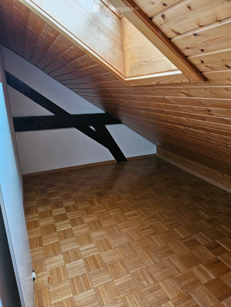Empty attic space with wooden herringbone flooring and exposed wooden beams on the ceiling. The walls are painted white, and the room has a slanted ceiling typical of an attic.