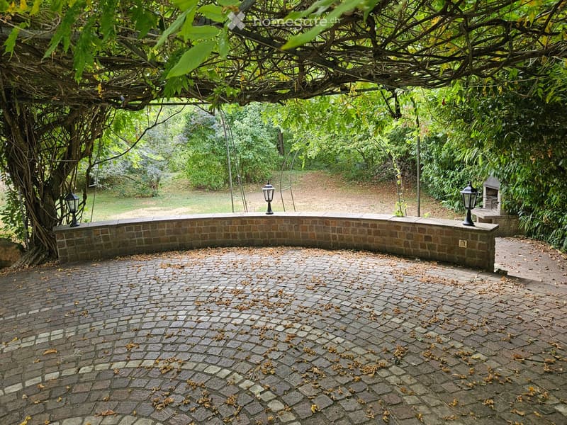 A paved outdoor area with a brick wall, lanterns, and a tree-covered pathway leading to a grassy area.