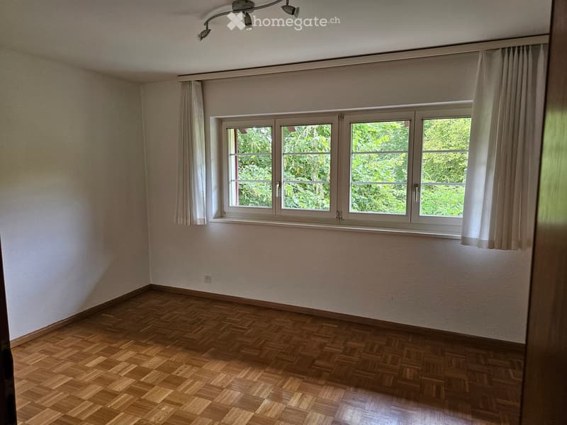 Empty room with wooden flooring, white walls, large window with white curtains, and a ceiling light fixture.