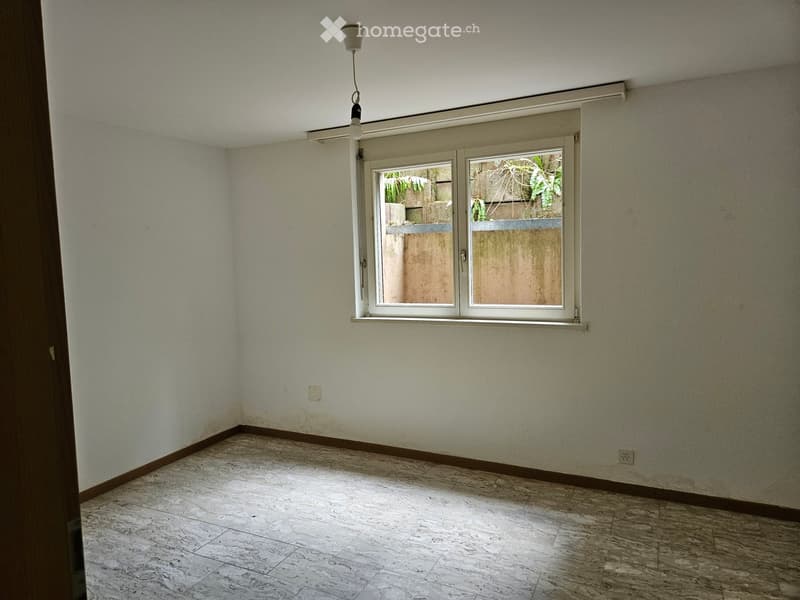Empty room with light-colored walls, marble flooring, and a single window with a view of a garden area.