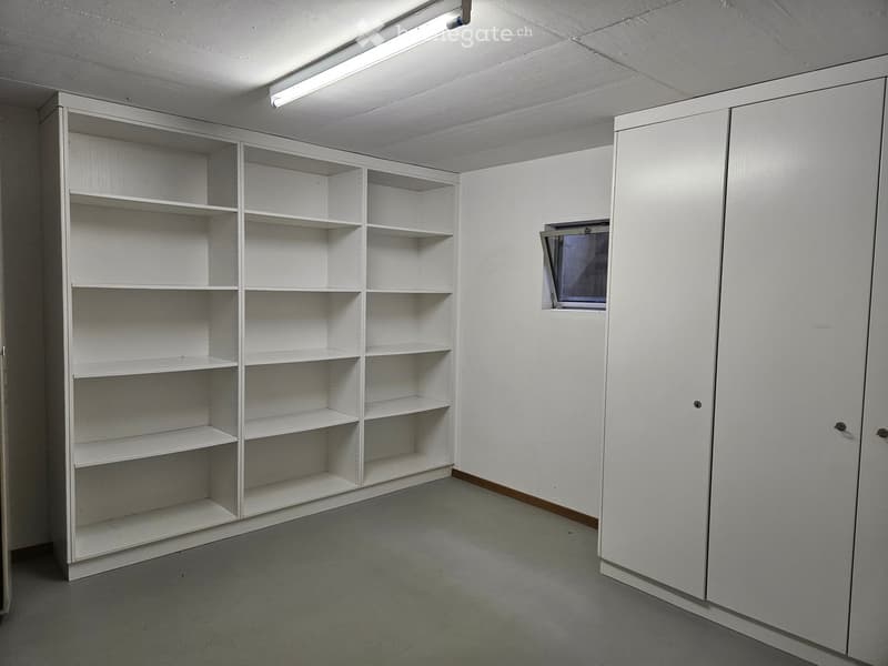 Empty room with white shelving units and built-in cabinets, featuring a small window and fluorescent lighting.