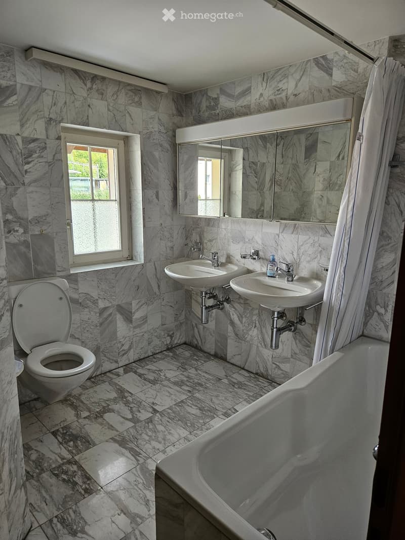 Bathroom with marble tiles, a toilet, a bathtub, two sinks, and a large mirror above the sinks.