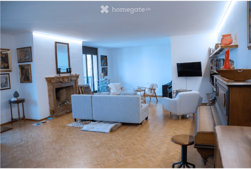Bright living room with wooden herringbone flooring, fireplace, large windows, balcony access, sofa, armchair, coffee table, TV, and decorative items.
