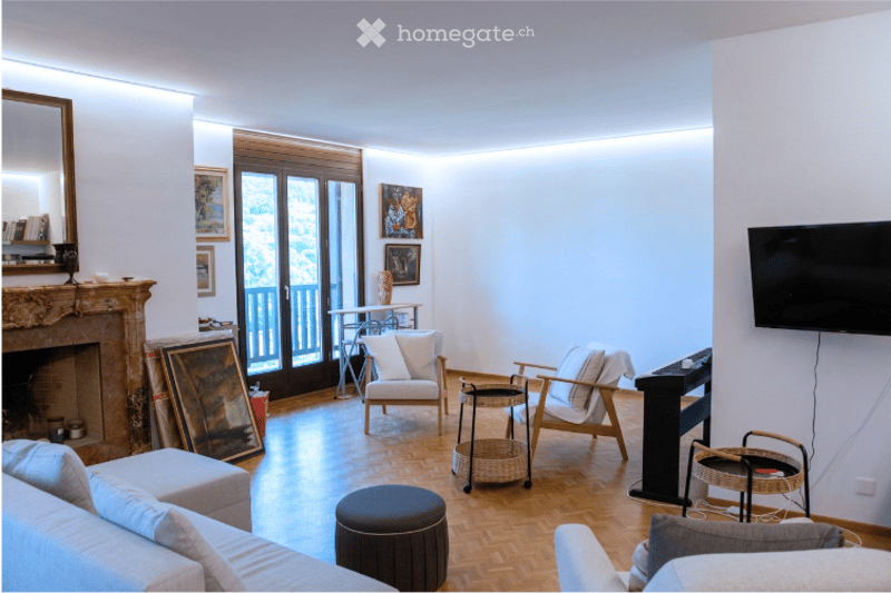 Bright living room with wooden flooring, white walls, fireplace, large windows, and balcony access. Features a sofa, ottoman, chairs, coffee table, and television.