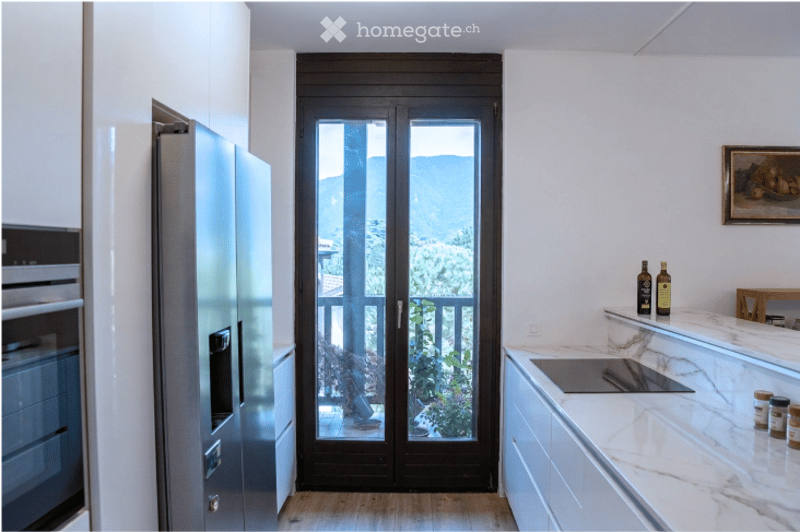 Modern kitchen with white cabinets, marble countertops, stainless steel appliances, and a large glass door leading to a balcony with a scenic mountain view.