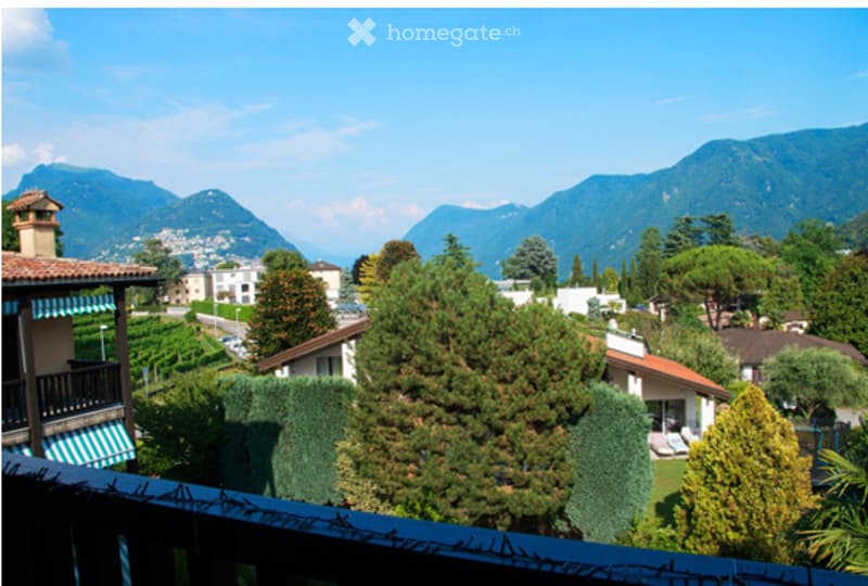 Balcony overlooking a scenic view of a town and surrounding mountains, with trees and houses below.