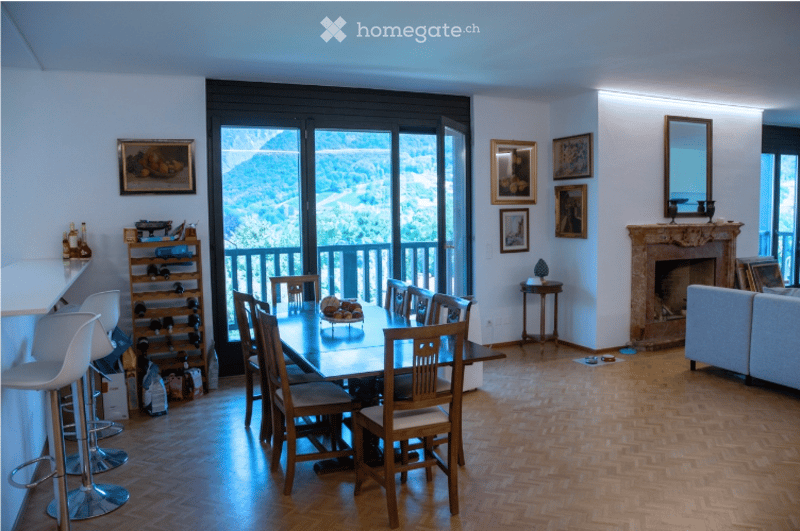 Open-plan living and dining area with wooden flooring, large windows offering a scenic view, a dining table with chairs, a sofa, a wine rack, and a fireplace with a mirror above it.