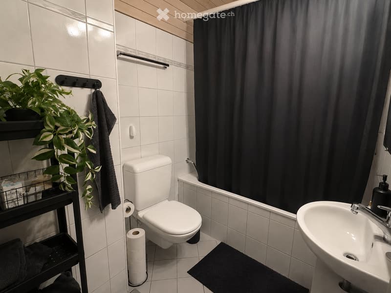 Modern bathroom with white tiles, a toilet, a sink, a bathtub with a dark curtain, a towel on a rack, a plant on a shelf, and a soap dispenser on the sink.