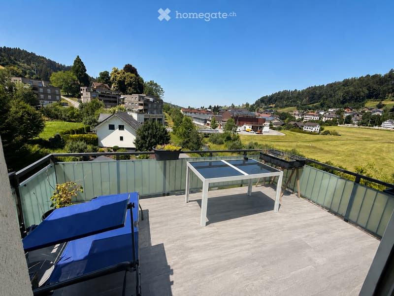 Balcony with a modern glass railing, a white table, a blue lounge chair, and potted plants, overlooking a green landscape with houses and hills.