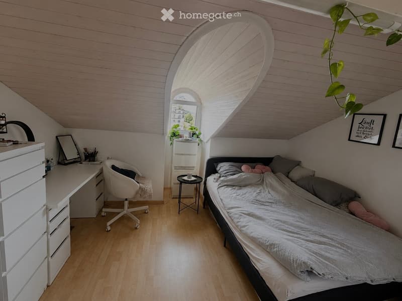 Cozy attic bedroom with slanted wooden ceilings, a bed with pillows and blankets, a white desk with a chair, a small round stool, and a framed picture on the wall.