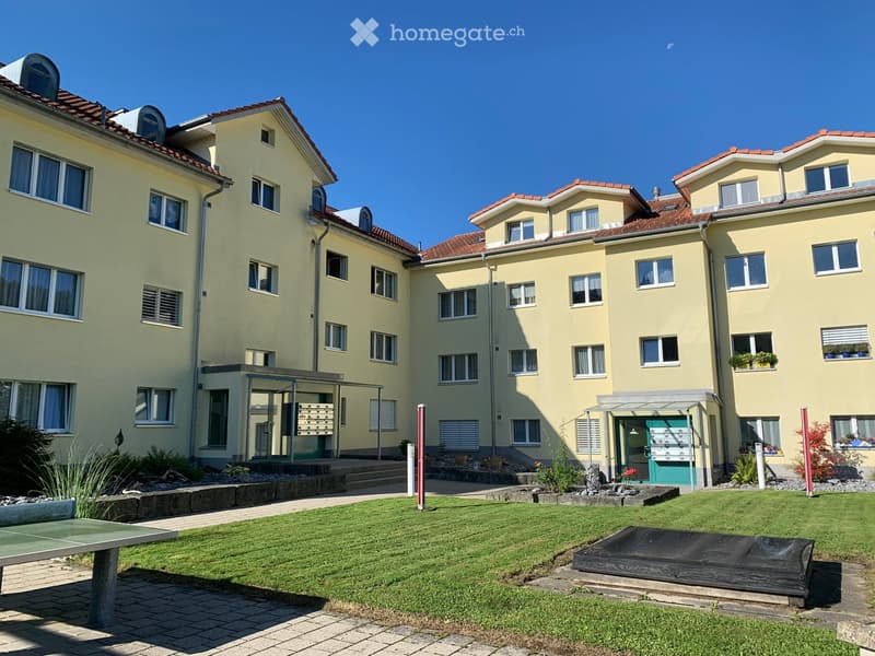 A multi-story residential building with yellow walls, red-tiled roofs, and multiple windows. The building features a well-maintained lawn, a ping pong table, and a small wooden platform in the foreground.