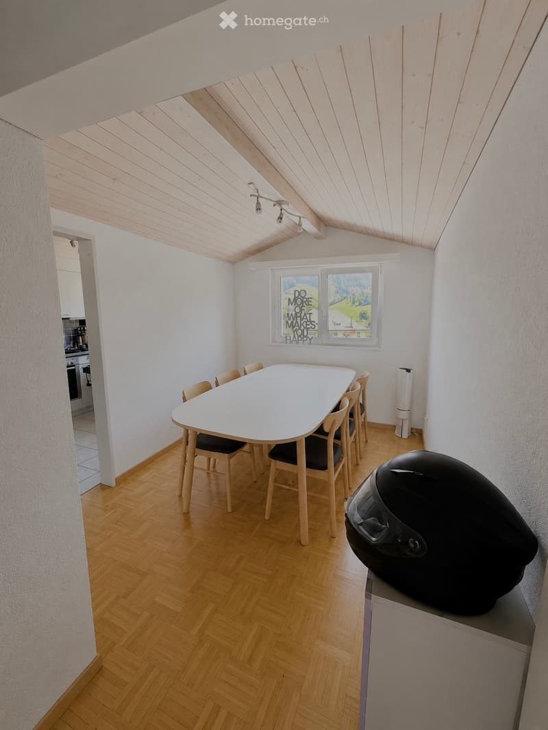 Bright dining area with a white table and six chairs, wooden flooring, and a large window with a motivational poster. A black helmet is placed on a surface in the foreground.