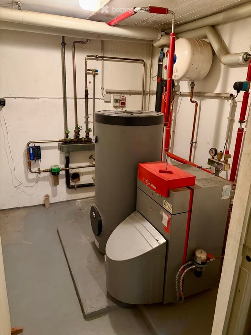 Utility room with a water heater, boiler, and various pipes and valves. The room has white walls and a concrete floor.